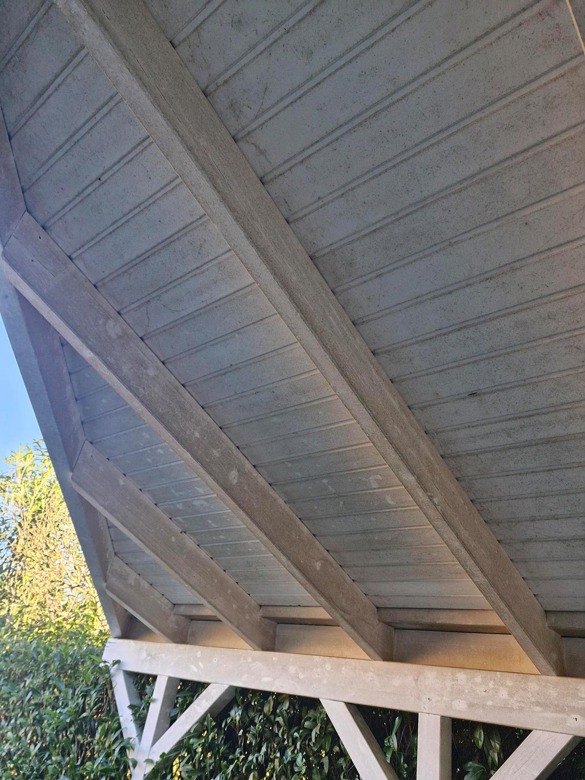 Ein hölzernes Dach mit sichtbaren Balken und grauen Schindeln über grünem Gebüsch