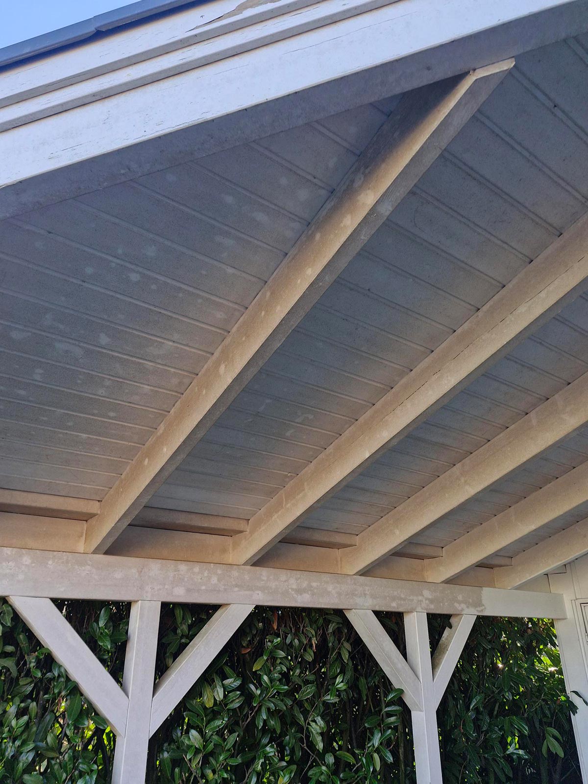 Ein hölzernes Terrassendach in hellem Beige vor grünen Blättern im Sonnenlicht