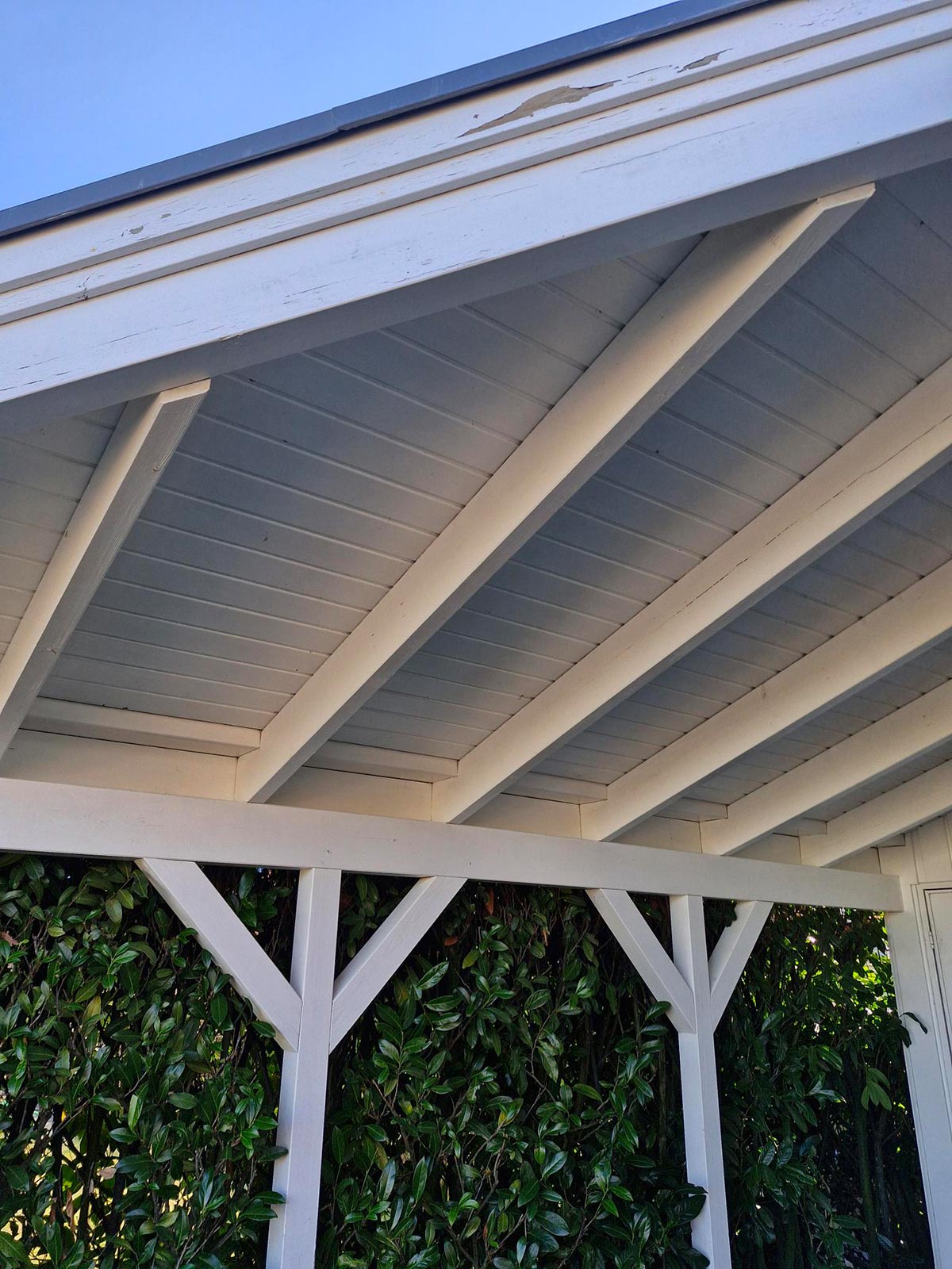 Überdachter weißer Holzunterstand mit sichtbaren Balken vor grüner Hecke und blauem Himmel