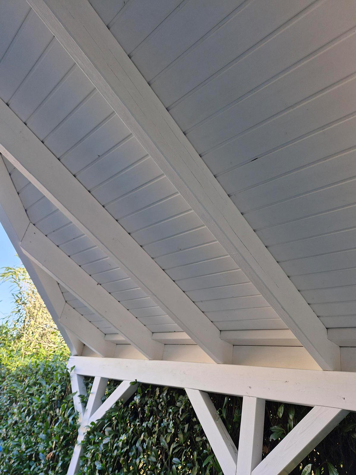 Ein weiß gestrichenes Holzdach mit schrägen Balken und grünen Büschen darunter