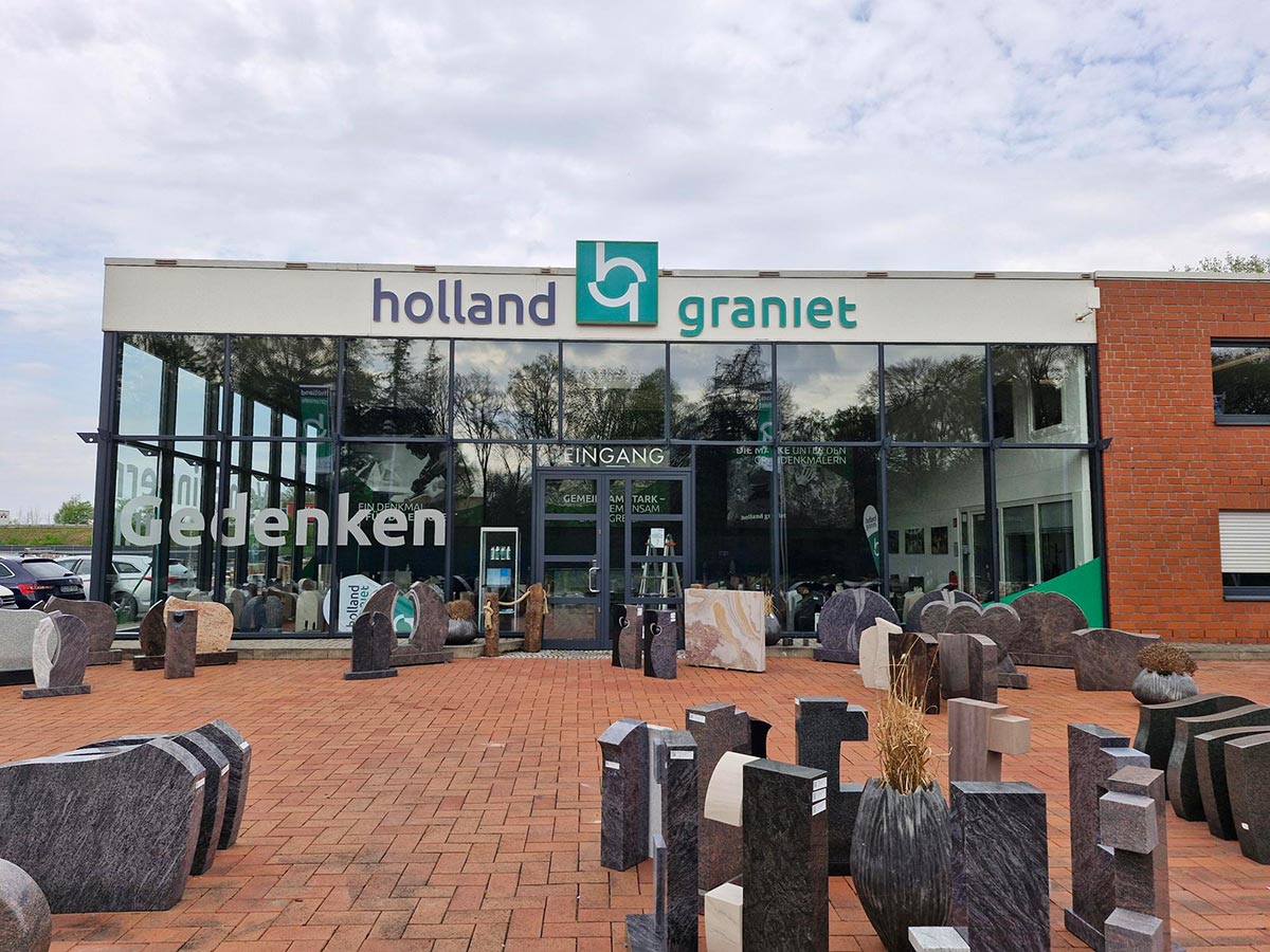 Vor einem modernen Gebäude mit Glasfront stehen verschiedenfarbige Grabsteine aus Granit