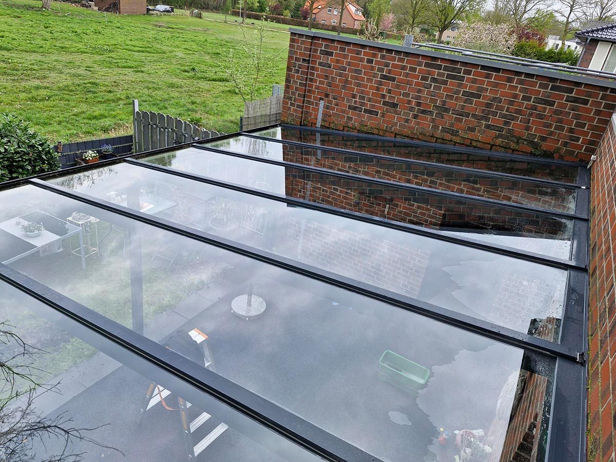 Glasüberdachung mit schwarzen Rahmen über einer Terrasse neben einer roten Backsteinwand