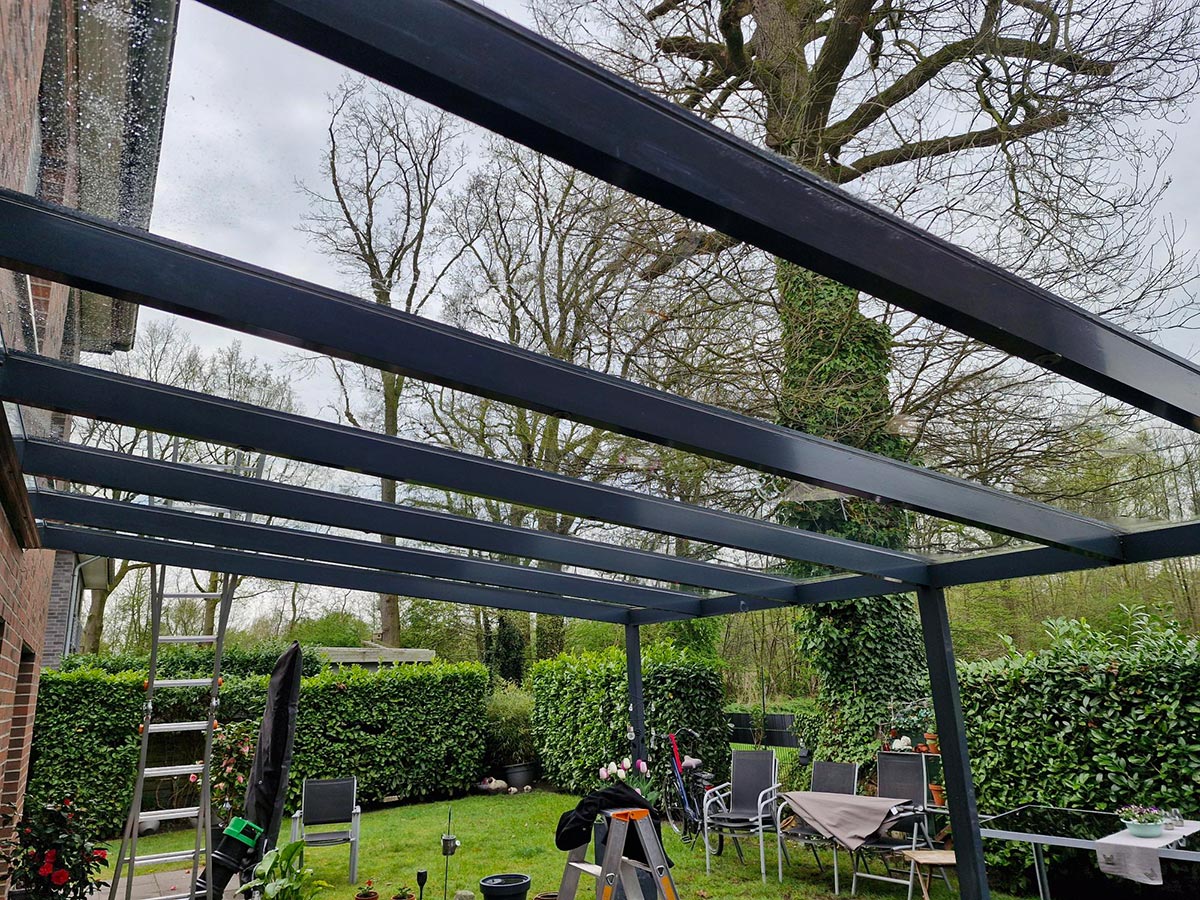 Metallüberdachung im Garten mit grauen Stützen, umgeben von grüner Hecke und Gartenmöbeln