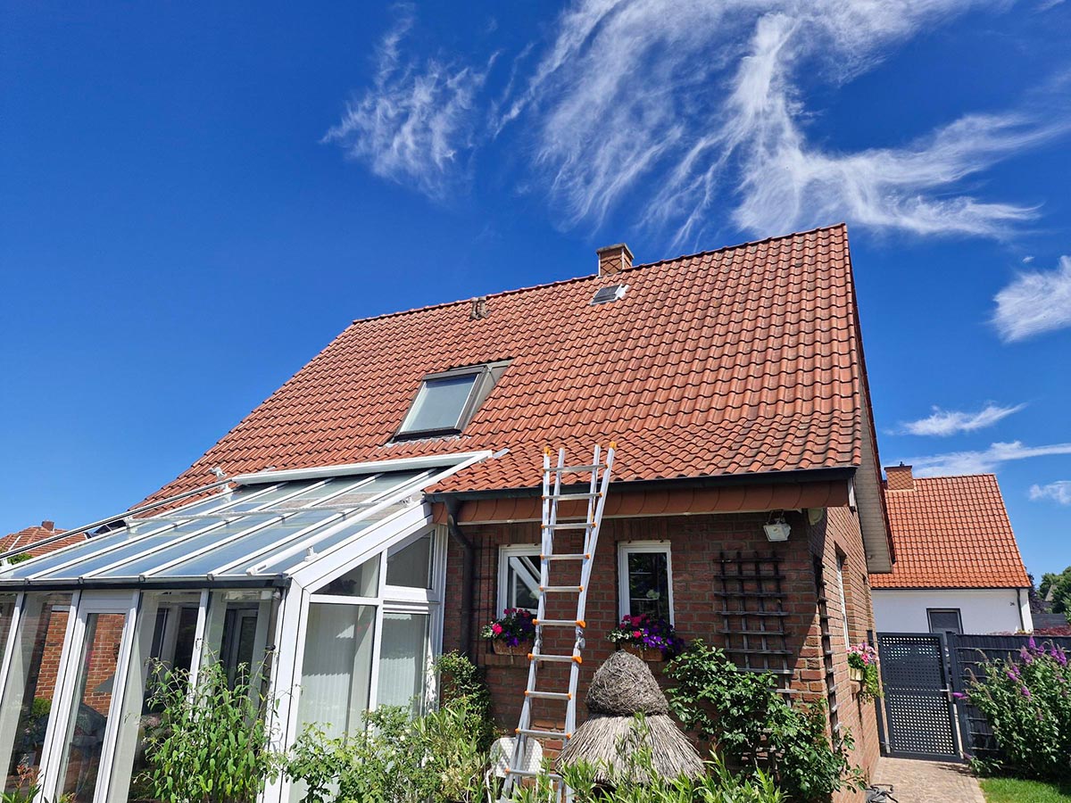Ein rotes Ziegeldachhaus mit Leiter, Wintergarten und blühenden Blumen vor blauem Himmel