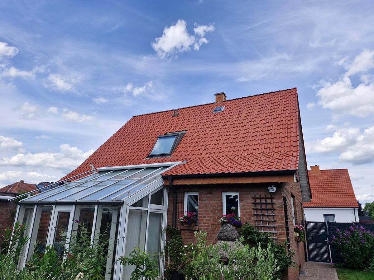 Ein rotes Ziegeldachhaus mit Wintergarten und vielen bunten Blumen vor blauem Himmel