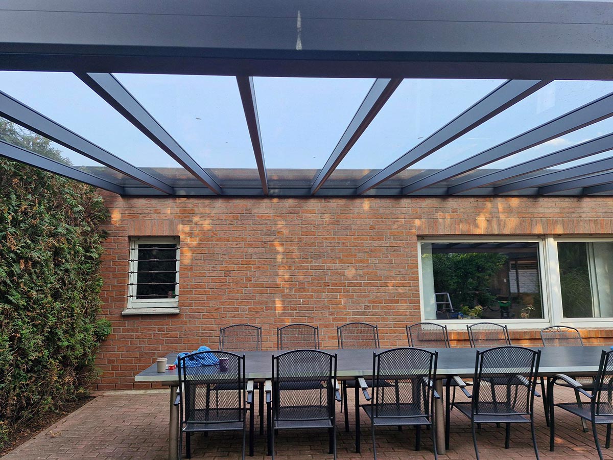 Mehrere schwarze Stühle stehen an einem langen dunklen Tisch unter einem Glasdach auf einer Terrasse mit Backsteinwand und großen Fenstern
